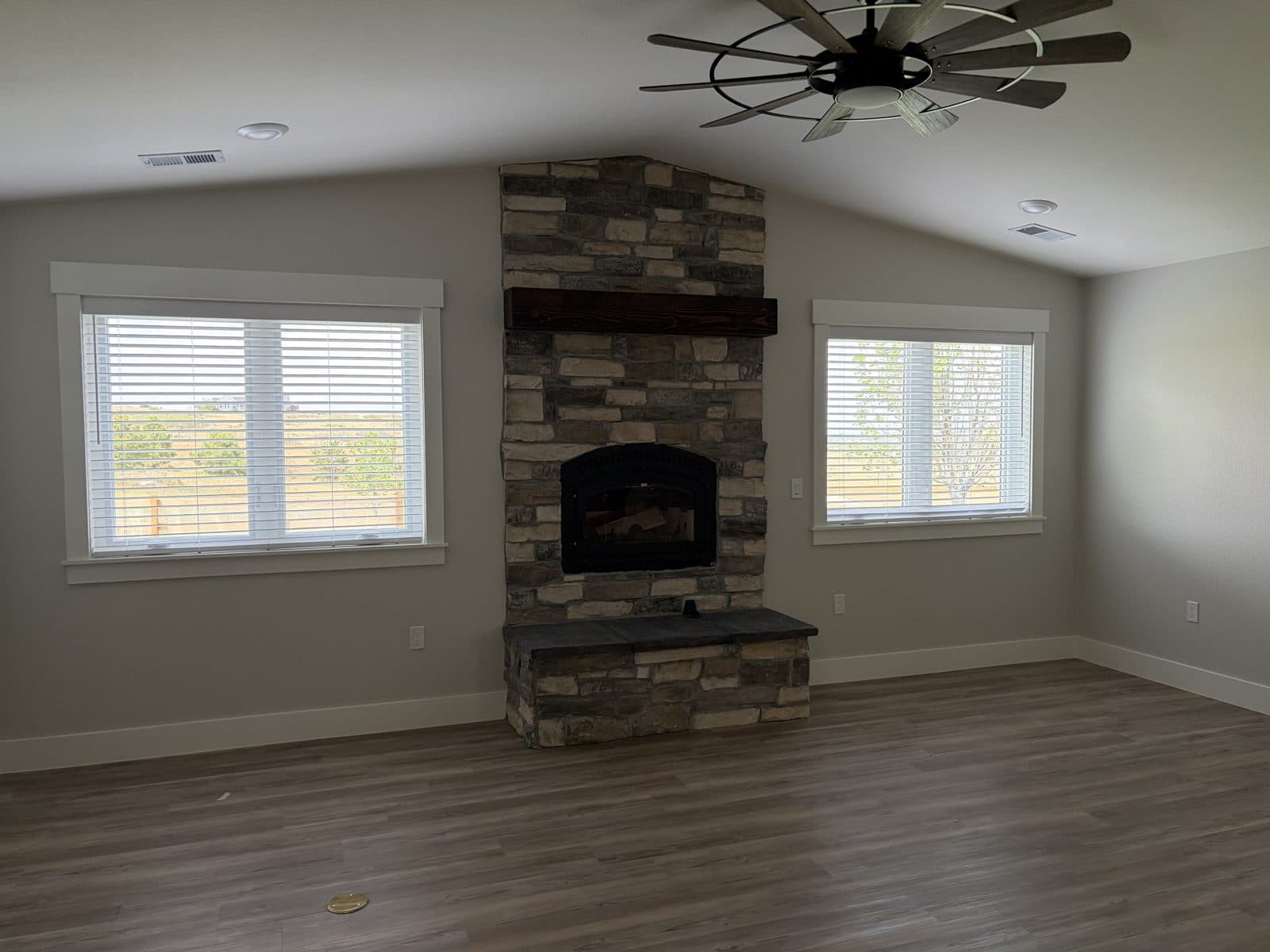 Great room with a stone fireplace and white blinds on the windows either side