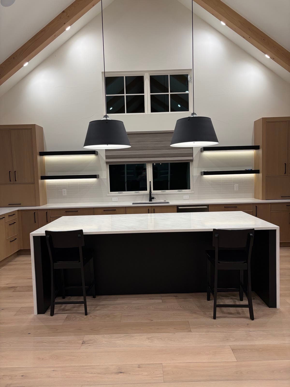 A Roman shade above the sink in a vaulted white-oak kitchen at night